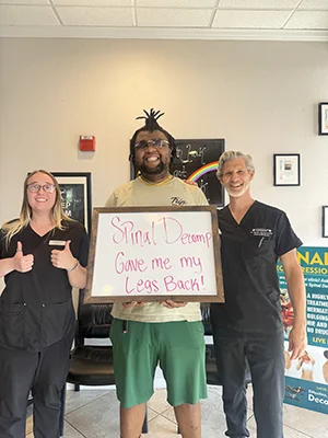 Patient with chiropractic staff holding a sign that reads, “Spinal Decomp gave me my legs back!”