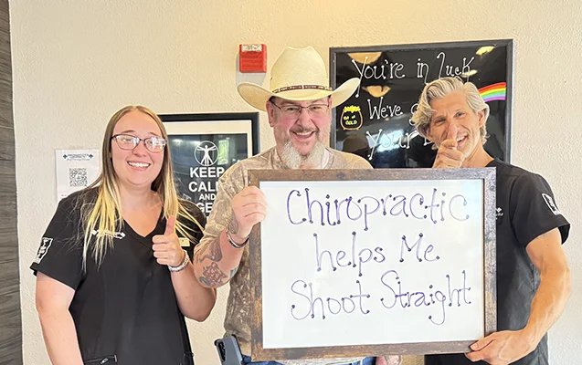 Patient with chiropractic staff holding a sign that reads, “Chiropractic helps me shoot straight.”