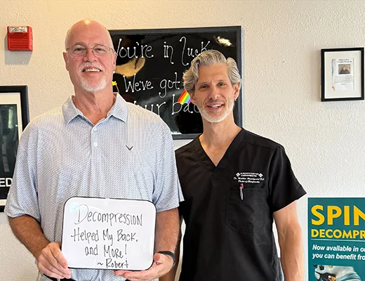 Patient with chiropractor holding a sign that reads, “Decompression helped my back, and more! – Robert”