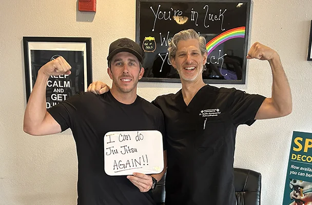 Patient and chiropractor flexing their arms while holding a sign that reads, “I can do Jiu Jitsu AGAIN!!”